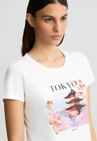 Valge puuvillane T-särk, millel on värvikas Tokyo graafika, kus on kujutatud pagoodi, kirsiõisi ja sõna "TOKYO" musta tekstiga.
