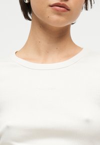 Γυναίκα που φοράει μικρούς ασημένιους κρίκους και λευκό ριμπ καλοκαιρινό τοπ Calvin Klein, εμφανιζόμενη από τον λαιμό μέχρι το στήθος μπροστά σε απλό φόντο.