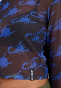 Črna mrežasta majica z modrim vzorcem škorpijona. Ima kratko zasnovo z blagovno oznako na robu, kar poudarja edinstven vzorec.