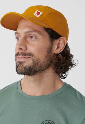 Uomo con barba leggera che indossa un berretto giallo senape con logo rosso e una maglietta verde con scollo rotondo, che guarda leggermente verso sinistra.