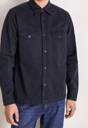 Homme portant une chemise noire à boutons avec deux poches poitrine, associée à un jean bleu, sur un fond clair uni.