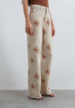 Pantalones beige de pierna ancha con bordado floral marrón usados por una persona de pie sobre suelo gris, combinados con top marrón y sandalias negras.