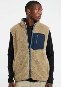 Beige Fleece-Weste mit hohem Kragen, Frontreißverschluss und navyblauen Taschenakzenten. Hat eine plüschige Textur und kontrastierende blaue Details.