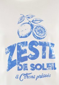 Vit t-shirt med blå vintage stil text: "ZESTE DE SOLEIL" tillsammans med illustrationer av hela och halverade citroner samt blad.