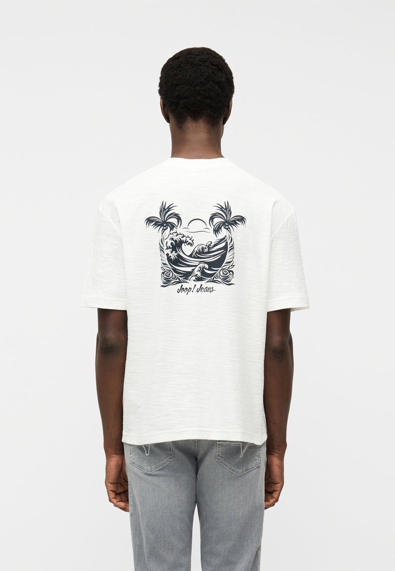 Μαύρο άτομο που φοράει λευκό T-shirt με μαύρο τροπικό κύμα και σχέδιο φοίνικα και γκρι τζιν, εμφανιζόμενο από πίσω.