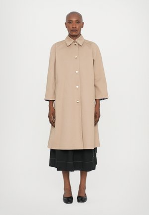 Cappotto beige di lunghezza al ginocchio con colletto a punta e chiusura frontale con bottoni, caratterizzato da una vestibilità comoda e maniche arrotolate a 3/4. Sotto, una gonna nera.