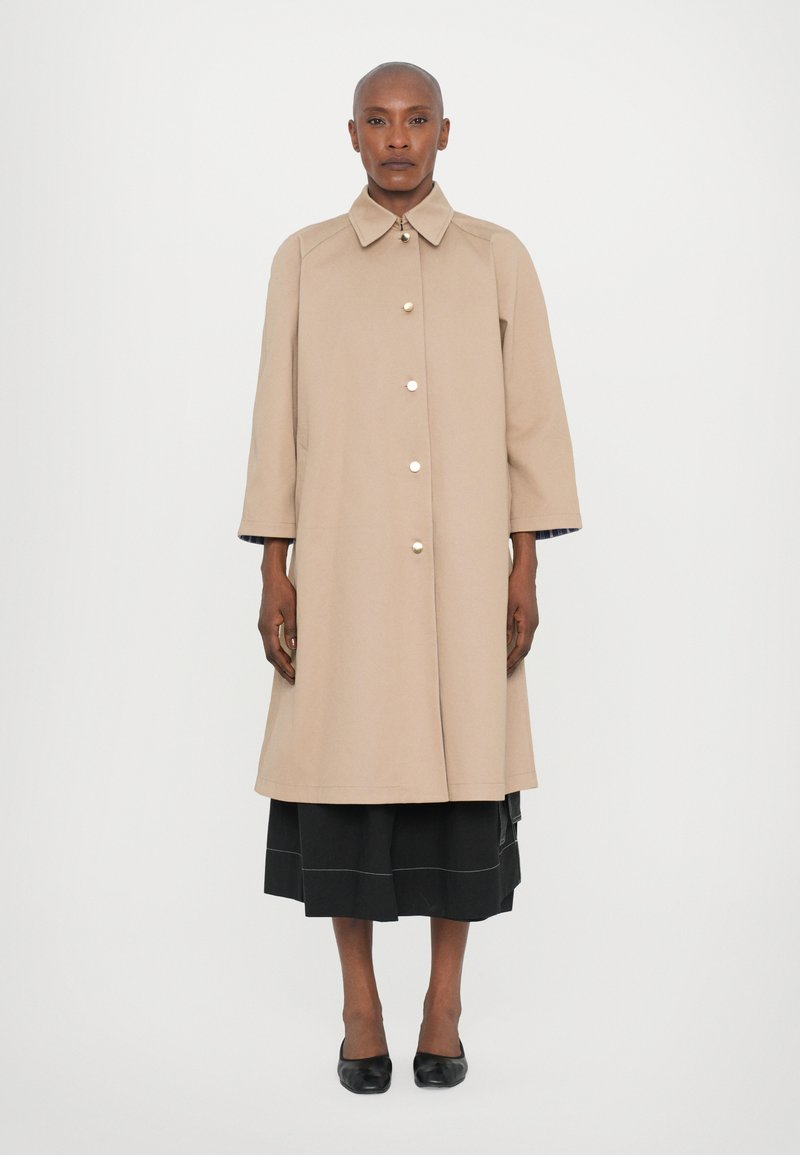 Cappotto beige di lunghezza al ginocchio con colletto a punta e chiusura frontale con bottoni, caratterizzato da una vestibilità comoda e maniche arrotolate a 3/4. Sotto, una gonna nera.