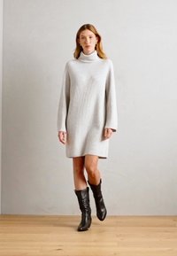 Svijetlosiva rebrasta pletena haljina s turtleneck izrezom i dugim rukavima, do sredine bedara, u kombinaciji s crnim čizmama do koljena. Glatka tekstura.