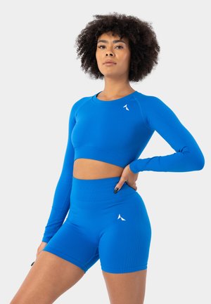 Kobieta z kręconymi włosami, ubrana w niebieski długi rękaw crop top i dopasowane szorty rowerowe z wysokim stanem, stoi z jedną ręką na biodrze.