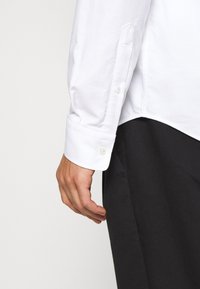 Main d'homme à côté du poignet d'une chemise blanche à manches longues et d'un pantalon noir, montrant les détails des boutons de la chemise et la texture du tissu.