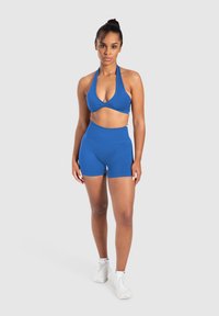 Vrouw die staat in een blauwe haltercrop top, hoge shorts en witte sportschoenen tegen een effen lichte achtergrond.