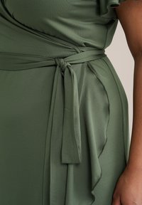 Rochie înfășurată dintr-un material neted de culoare verde măslin, având un cordon la talie legat, volane pe lateral și detalii de cusătură subtile.