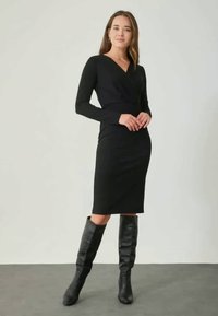 Svart omlottklänning med långa ärmar, knälång, figurnära silhuett. Matchas med knähöga svarta läderstövlar. Slät textur, minimalistisk design.