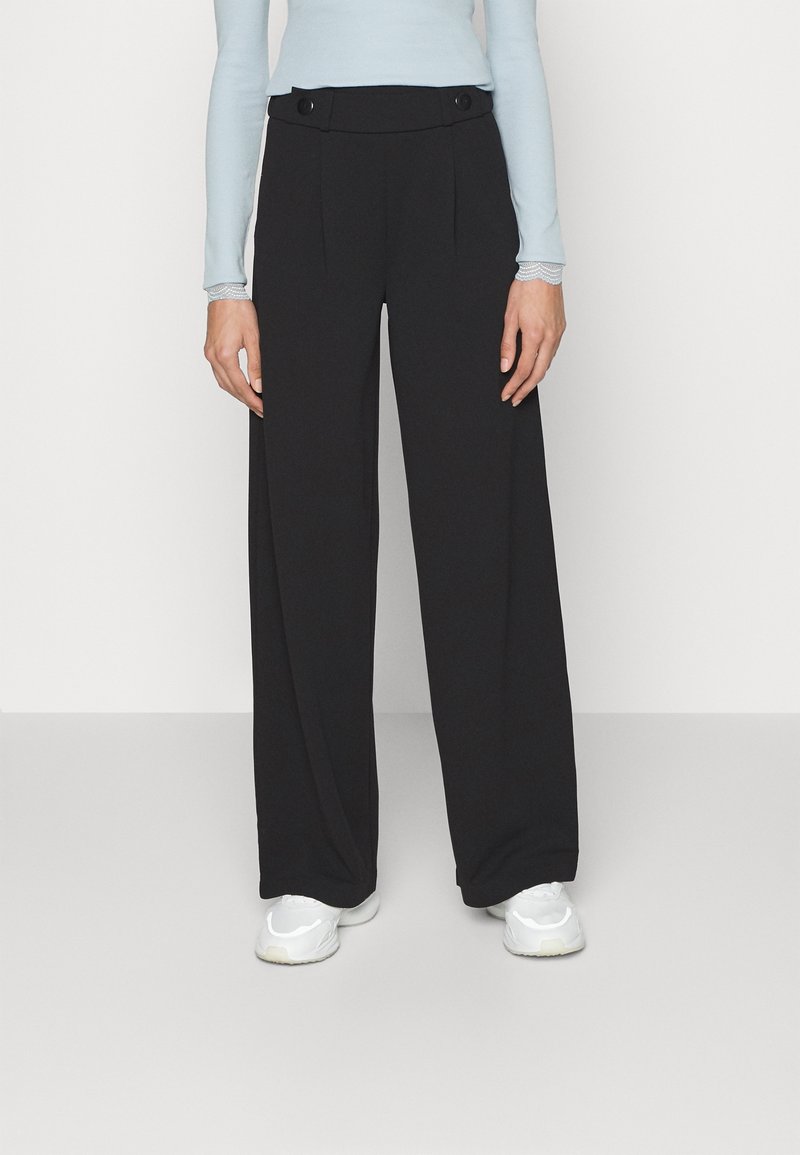 Personne portant un pantalon noir à jambes larges avec des boutons à la taille et des baskets blanches, associé à un haut bleu clair à manches longues.