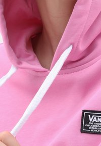 Sudadera rosa con cordones blancos, que presenta una etiqueta negra en la parte frontal con texto blanco. Material de algodón suave, diseño con capucha.