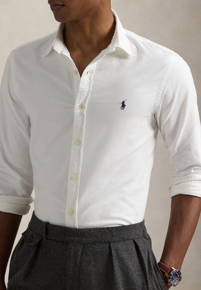 Hombre con camisa blanca de botones y mangas remangadas, pantalones plisados gris oscuro y un reloj de pulsera marrón con esfera oscura.