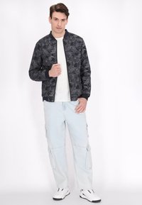 Zwarte bomberjack met wit bloemmotief, geribbelde mouwen en onderrand. Gecombineerd met lichtblauwe cargo pants en witte sneakers.