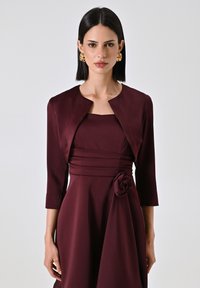 Bordeauxfarbenes Kleid mit tailliertem Oberteil, Dreiviertelärmeln und Taillierungsdetails mit einem Stoffrosenakzent; glattes, strukturiertes Material.
