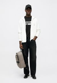 Άνδρας με μαύρο καπέλο, μαύρο μπλουζάκι Armani Exchange, λευκή φούτερ ζακέτα με φερμουάρ, μαύρα τζιν παντελόνια, μαύρα παπούτσια, κρατώντας γκρίζο σακίδιο από τη λαβή.