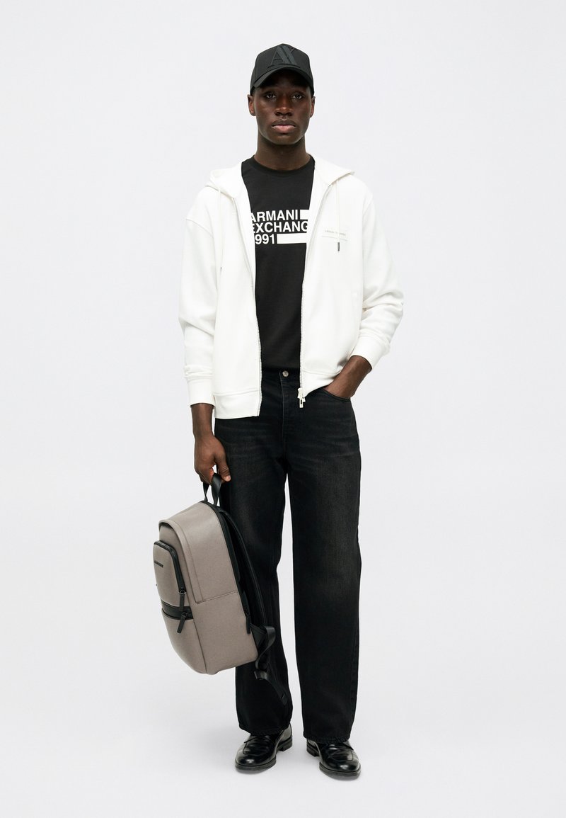 Άνδρας με μαύρο καπέλο, μαύρο μπλουζάκι Armani Exchange, λευκή φούτερ ζακέτα με φερμουάρ, μαύρα τζιν παντελόνια, μαύρα παπούτσια, κρατώντας γκρίζο σακίδιο από τη λαβή.