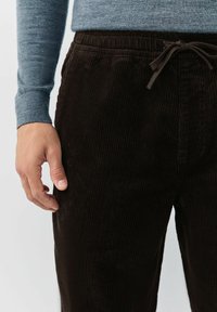 Man wearing dark brown corduroy pants with a drawstring waist and a blue long-sleeve shirt, hand relaxed by side.