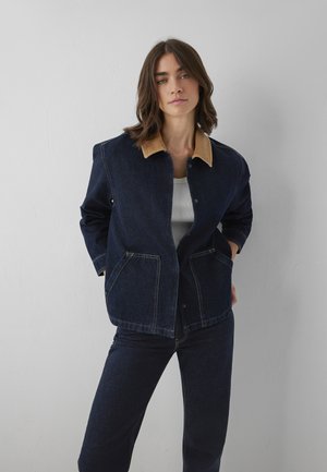 Femme portant une veste en jean bleu foncé avec un col beige et un jean assorti, debout les mains dans les poches de la veste, devant un mur clair uni.