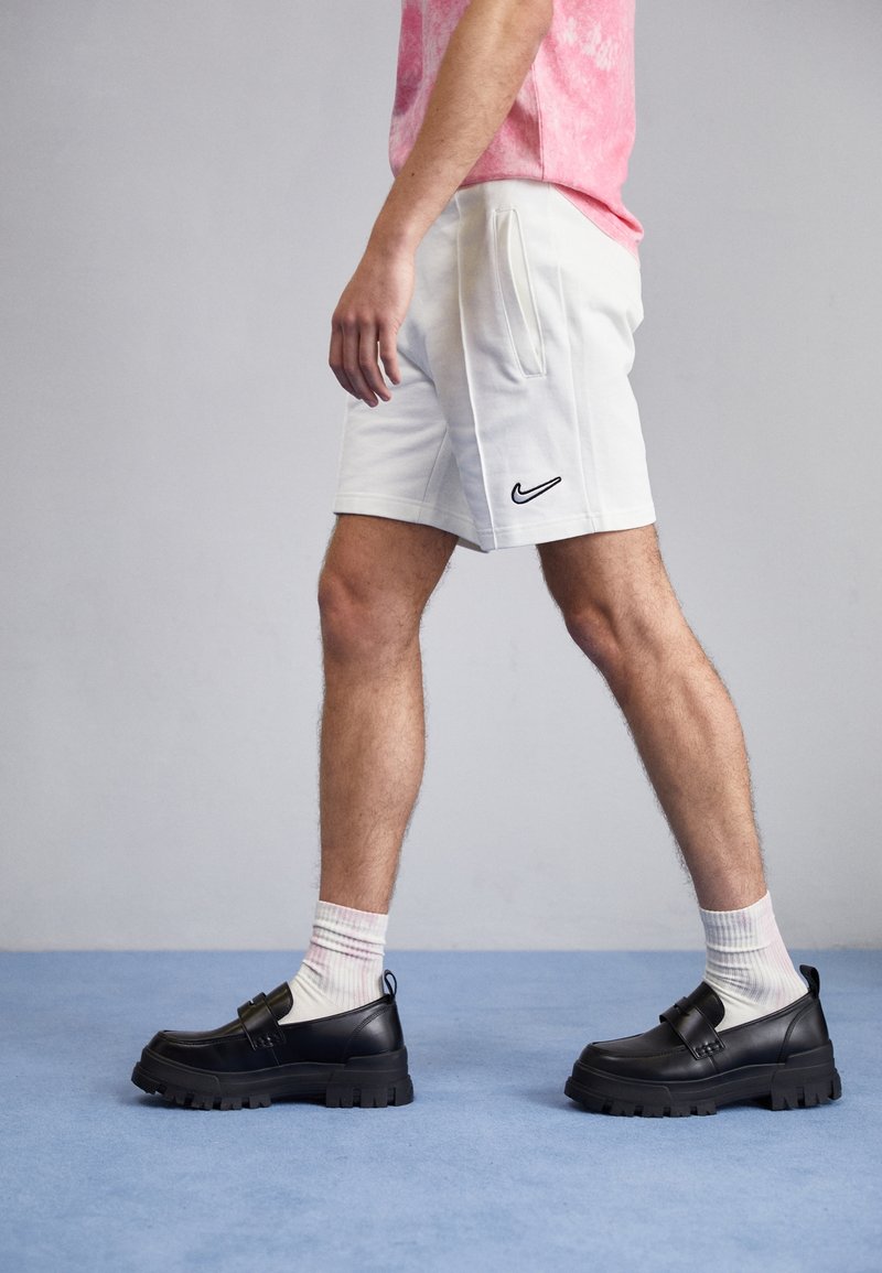 Black leather shoes with a chunky sole and strap detail, paired with white shorts and pink patterned t-shirt. Background is light gray.