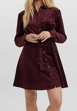 Kobieta w bordowej sukience z prążkowanego sztruksu z długimi rękawami, zapinanej na guziki, z paskiem wiązanym w talii, stojąca na jednolitym tle.