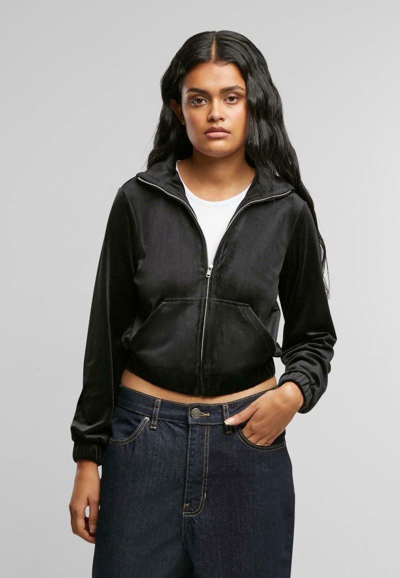 Svart kort zip-up hettegenser laget av myk stoff med frontlommer og elastiske mansjetter, kombinert med mørkeblå jeans med gyldne sømmer.