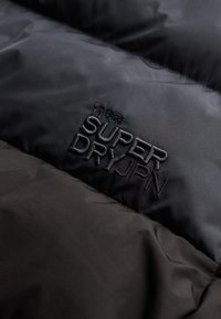 Zbliżenie na czarny materiał wyściełany, z haftowanym logo Superdry JPN oraz japońskimi znakami.