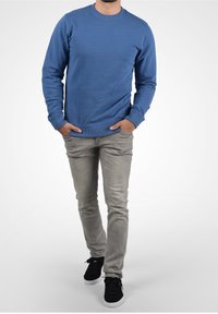 Niebieska bawełniana bluza z długimi rękawami, ściągaczami przy mankietach i dekolcie, w zestawieniu z jasnoszarymi jeansami i czarnymi trampkami. Prosty wzór, bez nadruków.