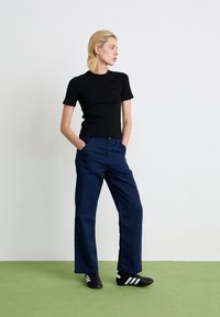 Jeune personne se tenant avec les mains dans les poches, portant une chemise noire à manches courtes, un pantalon large bleu marine et des baskets noires avec des rayures blanches.