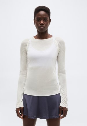 Rövid hajú nő fehér hosszú ujjú inget és tengerészkék szoknyát viselve, egy egyszerű világos háttér előtt áll.