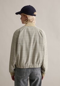 Καρό μπουφάν τύπου bomber σε μπεζ και ναυτικό μπλε, με ριμπ γιακά και ελαστική ζώνη. Συνδυασμένο με γκρι τζιν, σε θέα από πίσω.