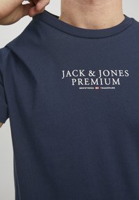 Tamno plava pamučna majica s bijelim printanim logotipom "JACK & JONES PREMIUM." Jednostavan dizajn s krušnim izrezom i kratkim rukavima.
