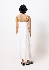 Weißes, besticktes Maxikleid mit dünnen Trägern und fließender Silhouette. Kombiniert mit schwarz-weißen gestreiften Schuhen.