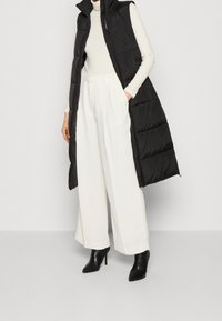 Svart lång vadderad väst med hög krage, bärs över en vit polotröja och vita vida byxor. Svarta spetsiga ankelstövlar fulländar outfiten.