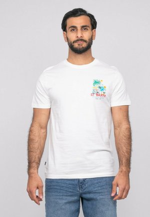 Homme aux cheveux foncés et à la barbe, portant un t-shirt blanc avec un motif coloré de la plage de St Barth sur la poitrine, associé à un jean bleu, debout contre un fond clair.
