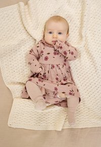 Babaruha lágy rózsaszín anyagból, virágmintával és fodros gallérral, világos bézs kötött harisnyával párosítva. Texturált krémszínű takarón rétegezve.