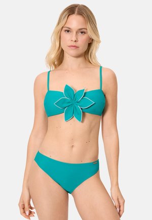 Mujer con bikini color verde azulado de tirantes delgados, con una gran adorno en forma de flor en el centro superior, posando frente a un fondo liso.