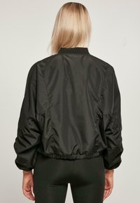 Black bomber jacket with a shiny, smooth texture. Features gathered sleeves and a ribbed neckline, designed with an elastic hem.