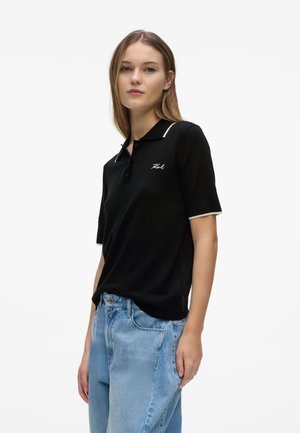 Ung kvinde iført en sort kortærmet polo med hvide kanter og lyseblå højtaljede jeans, stående mod en ensfarvet baggrund.