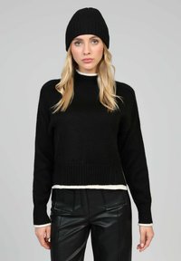 Pull en tricot noir à col montant et ourlet blanc, associé à un pantalon en cuir noir et un bonnet côtelé assorti, avec des manches longues.