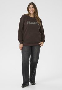 Felpa marrone con la scritta "FEMME" in caratteri decorativi, abbinata a jeans grigi scuri a zampa. Il tessuto sembra morbido; sfondo semplice.