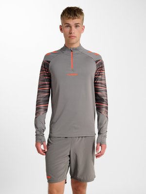 Jeune homme portant un haut de sport gris à manches longues avec des manches à motifs et un short assorti, debout devant un fond blanc uni.