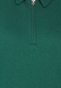 Polo shirt verde a coste con zip parziale e accessori in argento, colletto robusto e una texture liscia su tutto il tessuto.