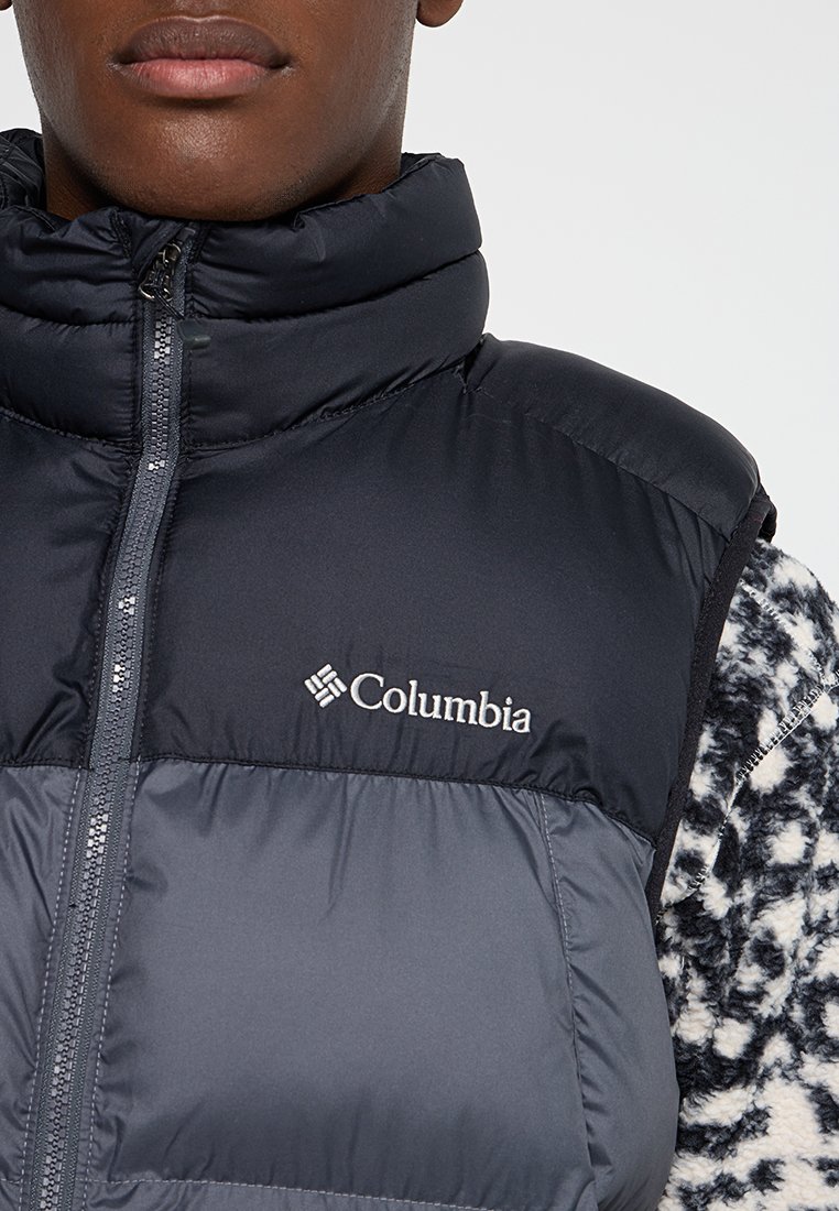 Kamizelka puchowa Columbia w ciemnoszarym kolorze, z wysokim kołnierzem, pełnym zamkiem błyskawicznym i logo. Materiał wydaje się gładki i izolowany, o eleganckim wyglądzie.