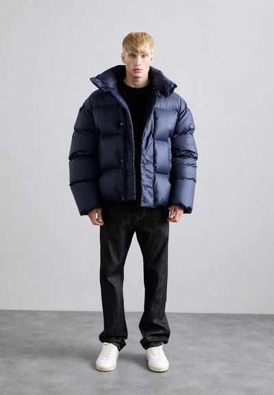 Ein junger Mann steht mit dem Gesicht nach vorne in einer marineblauen Steppjacke, schwarzen Jeans und weißen Sneakers vor einem schlichten grauen Hintergrund.