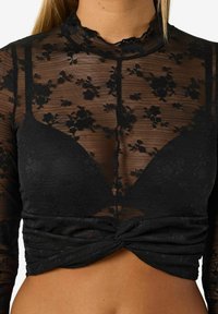 Fekete, hosszú ujjú, áttetsző virágmintás csipkéből készült felső, amelynek illeszkedő, megkötős eleje és magas nyaka van. A texturált felület kiemeli az anyagot.