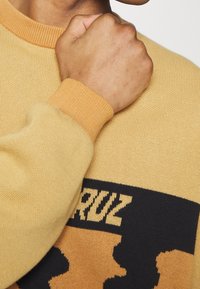 Žlutý pletený svetr s černým vzorem, s vroubkovaným kulatým výstřihem a manžetami. Textura látky je jemná a ukazuje polohu rukou.